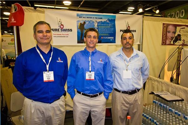 Three men in blue button-up shirts and lanyards stand side by side in front of a Pure Swiss banner showing a mountain scene, with bottled water on a table at right.