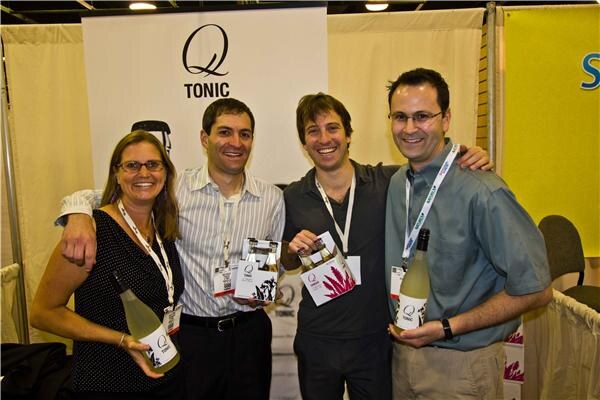 A woman and three men pose close together in front of a Q Tonic sign, holding bottles and a four-pack of Q Tonic, all smiling and wearing lanyards.