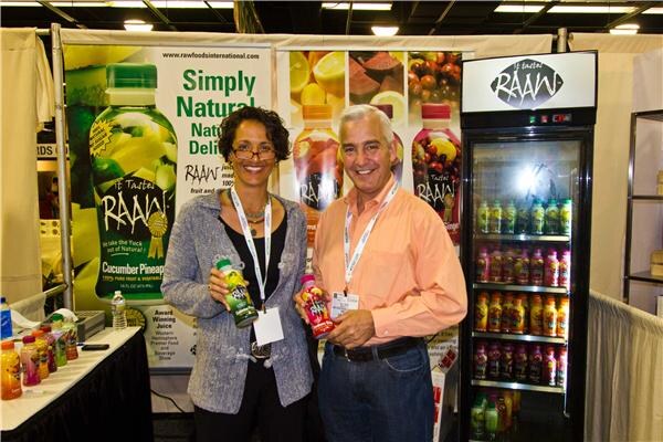 A woman and a man holding bottles of juice stand in a booth with Raaw banners and a lit Raaw refrigerator stocked with bottles; Raw Foods International appears on the signage.