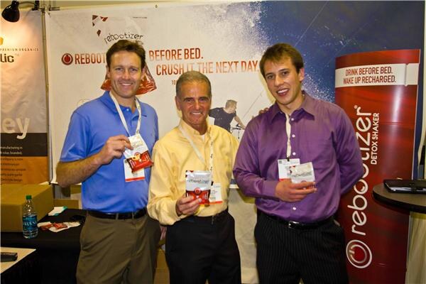 Three men hold small Rebootizer packets in front of a Rebootizer banner reading 'Before bed. Crush it the next day,' beside a red Rebootizer Detox Shaker display.