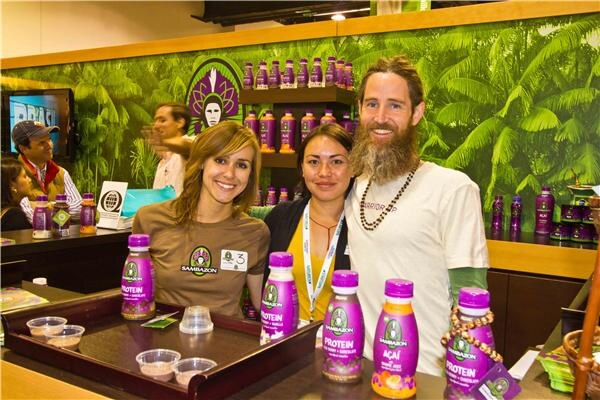 Two women and a bearded man smile behind a counter with Sambazon protein and açai bottles, in front of a jungle-themed backdrop with shelves of purple Sambazon bottles.