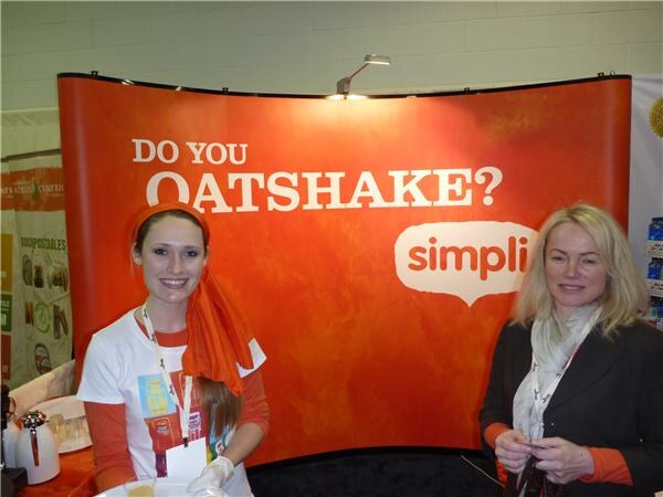A young woman in an orange head scarf and white shirt and a blonde woman in a dark jacket stand before an orange simpli display reading 'Do you oatshake?'