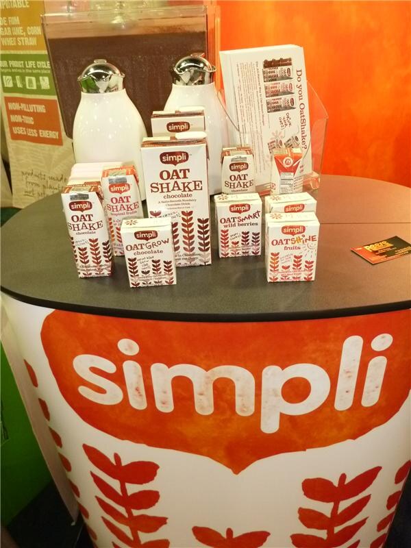 Display of simpli oat drink cartons, including Oat Shake, OatGrow, OatSmart and OatShine, on a black tabletop above an orange simpli-branded base, with two white dispensers behind.