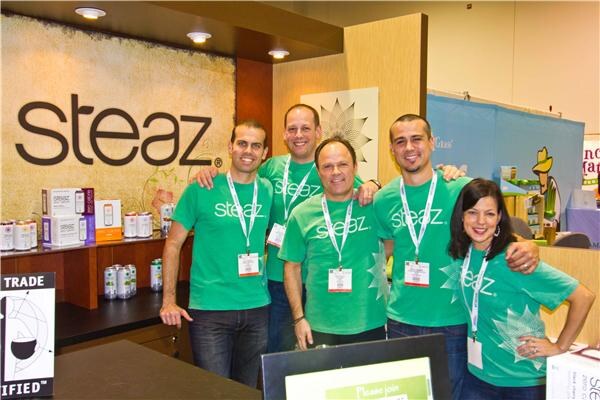 Five people in green Steaz T-shirts and badge lanyards stand together smiling in front of a Steaz sign, with product cans and boxes on a counter to the left.
