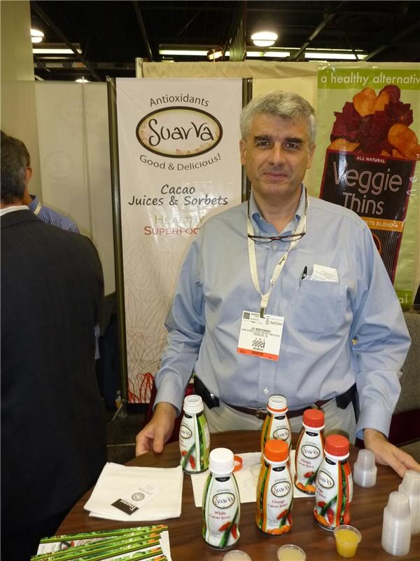 A gray-haired man in a light blue shirt and badge stands behind a table of Suavva cacao juice bottles, with a Suavva banner and a Veggie Thins banner behind him.