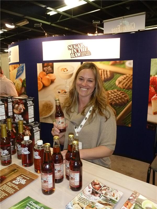 A smiling woman with long blonde hair holds a bottle of Sugar in the Raw syrup at a table of bottles and boxes, in front of a Sugar in the Raw backdrop with food photos.
