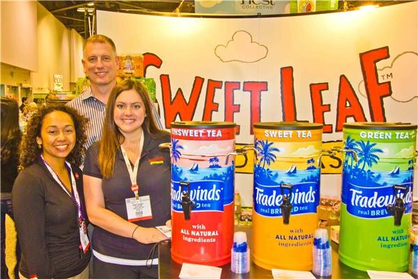 Two women and a man smile behind three Tradewinds tea dispensers labeled Unsweet Tea, Sweet Tea and Green Tea, beneath a Sweet Leaf sign.