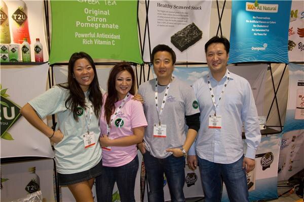 Two women and two men wearing badges pose together in a booth with T2U green tea banners and a Dongwon 100% All Natural seaweed snack banner behind them.