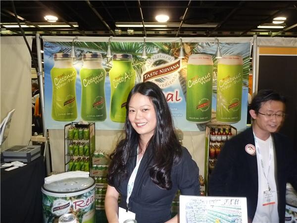 A woman with long dark hair stands in front of a Taste Nirvana coconut water banner; a man in glasses stands at right, with product displays and a cooler nearby.