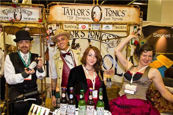 Four people in vintage-style costumes pose at a Taylor's Tonics booth, with bottles of botanical soda on the counter and a Taylor's Tonics sign overhead.