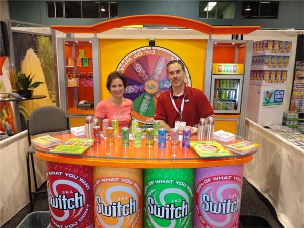 A woman and a man sit behind an orange counter topped with The Switch beverage cans, with a prize wheel behind them and a base decorated with large Switch can graphics.