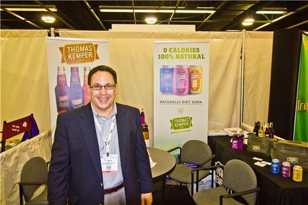 A smiling man in glasses, a dark blazer and a lanyard stands in the Thomas Kemper booth beside two banners for Thomas Kemper soda, with a table of bottles and cans behind him.