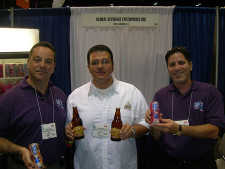 Three men with name badges pose at the Global Beverage Enterprises Inc booth. Two hold cans and one in a white shirt holds two brown bottles, in front of a white and blue curtained wall.