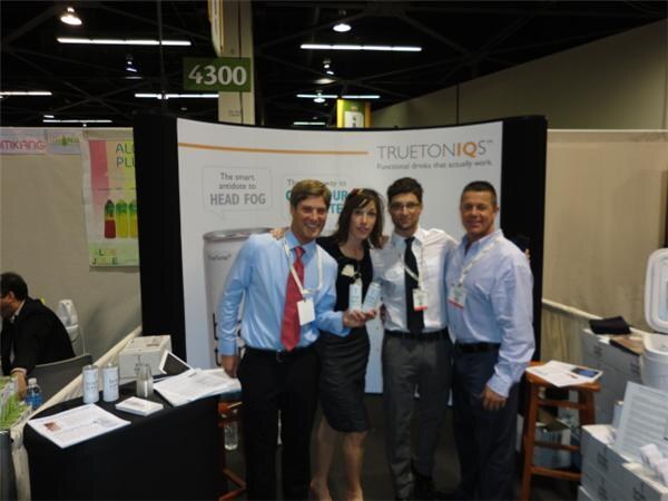 Three men and a woman stand together at a TruetonIQs booth, with a banner about head fog behind them and papers and cans on the tables.