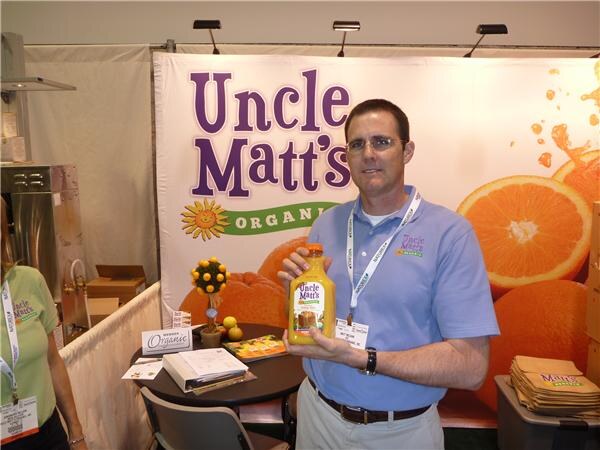 A man in a blue polo and lanyard holds a bottle of Uncle Matt's Organic orange juice in front of a large Uncle Matt's Organic banner with orange slices; part of another person is at left.