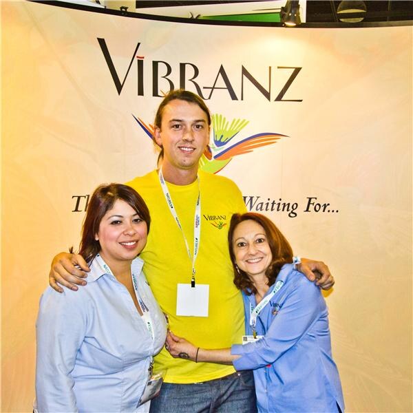 A tall man in a yellow Vibranz t-shirt stands with his arms around two women in light blue shirts, all smiling, in front of a Vibranz banner with a colorful bird logo.