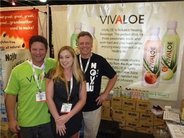 Two men and a woman with badges stand in a booth with a Vivaloe aloe vera drink banner and stacked NoYu boxes behind them; one man wears a green shirt, the other a black NoYu shirt.