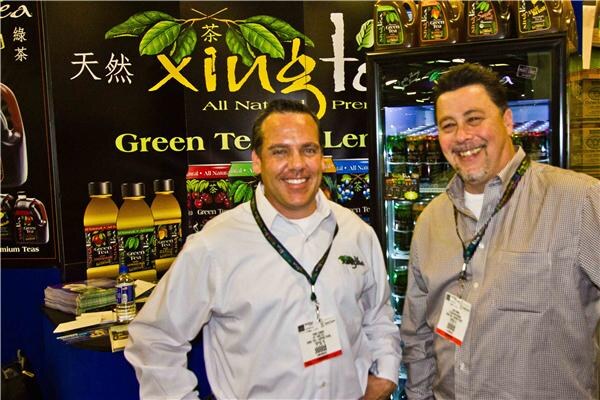 Two smiling men wearing lanyards and button-up shirts stand in front of a Xing Tea display of green tea bottles and a lit refrigerated cooler.