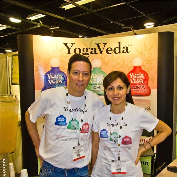 A man and a woman in white YogaVeda t-shirts with badges stand in front of a YogaVeda display showing blue, green and red bottles.