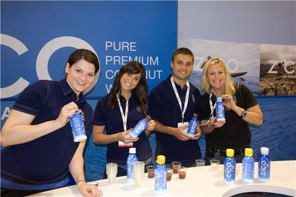Four smiling people in dark shirts hold bottles of Zico coconut water at a counter with more bottles and small sample cups, in front of a blue Zico backdrop.