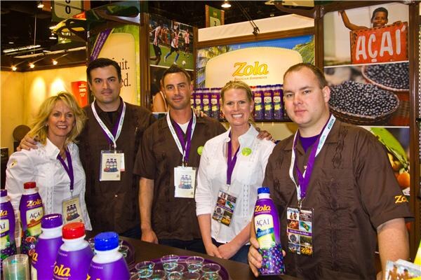 Five people in brown and white shirts with lanyards stand together at the Zola booth; the man at right holds a bottle of Zola açai juice, with purple bottles on the counter.