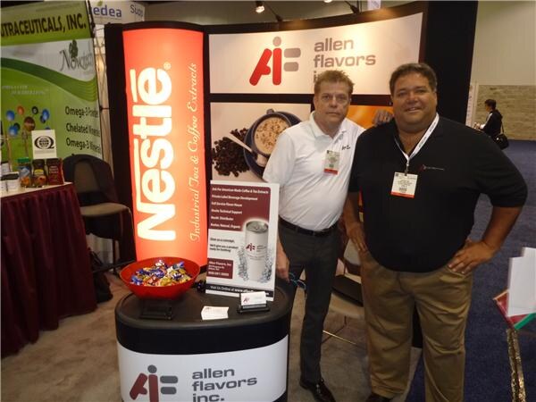 Two men with badges stand at an Allen Flavors booth with a Nestlé Industrial Tea & Coffee Extracts display and a bowl of wrapped candy on the counter.
