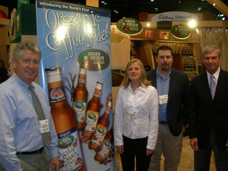 Two men and a woman stand beside a Green Mountain Coffee Roasters banner reading Organic Coffee Soda, with a fourth man at right, in an exhibit hall.