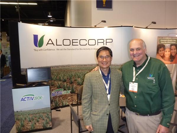 Two smiling men with badges stand in front of an Aloecorp backdrop showing an aloe field, beside an ActivAloe display.