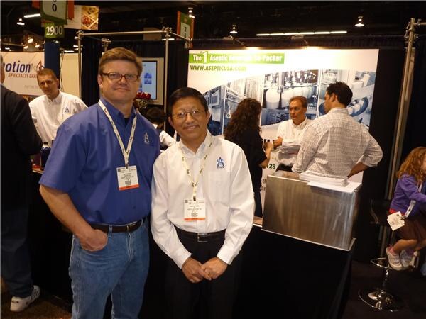 Two men in blue and white shirts with lanyards stand in front of an Aseptic USA booth graphic while other people stand behind a counter.