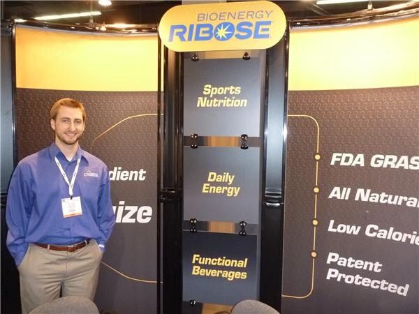 A smiling man in a blue shirt and lanyard stands beside a Bioenergy Ribose display listing sports nutrition, daily energy and functional beverages.