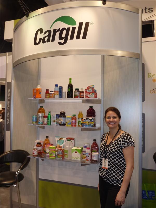 A smiling woman in a black and white top stands beside a Cargill display with shelves of bottled and boxed products, including Truvia.