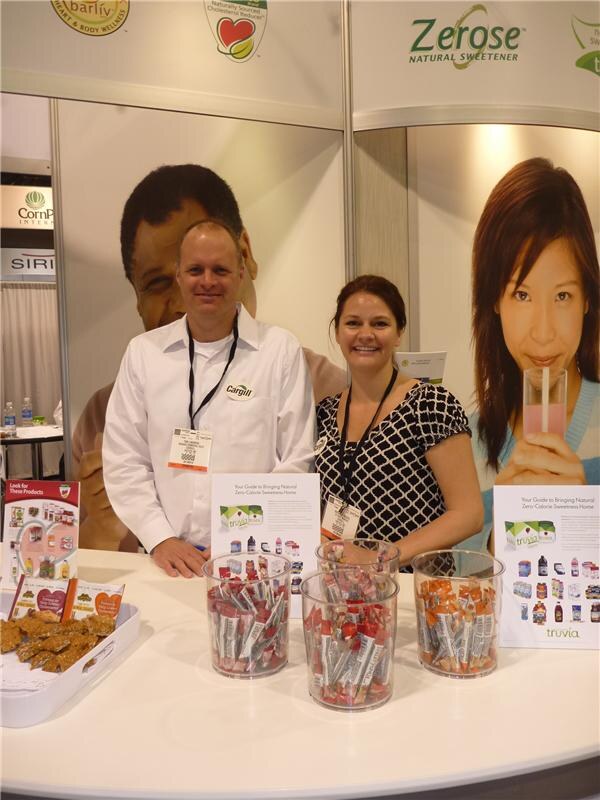 A man in a white shirt and a woman in a patterned top stand behind a counter with Truvia sweetener samples and brochures, under a Zerose natural sweetener sign.