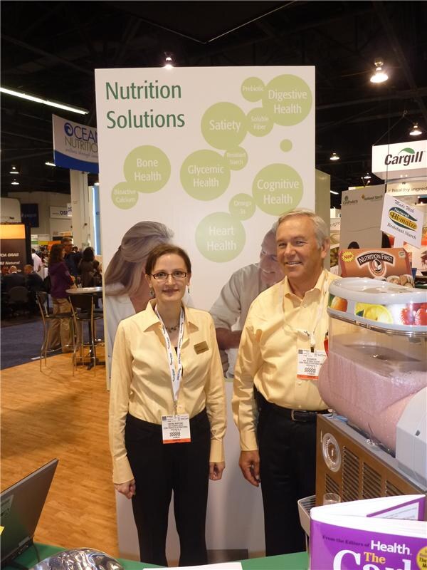 A woman and a man in yellow shirts with badges stand before a Nutrition Solutions panel; Hi-maize and Nutrition First products, plus Cargill and Ocean Nutrition signs, are visible.
