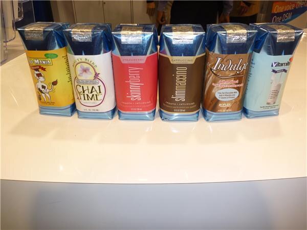 Six blue-and-silver aseptic drink cartons stand in a row on a white counter. Labels include Chai Time, Skinnyberry, Slimmacino, Indulge and Vitamite, plus one with a cartoon cow.