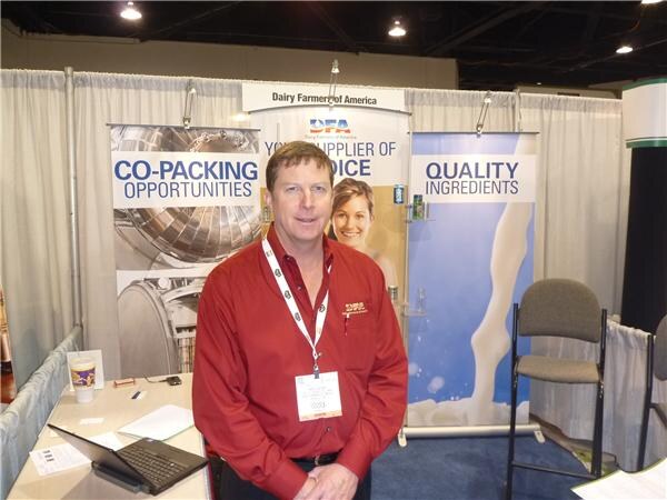 A man in a red shirt and lanyard stands in the Dairy Farmers of America booth, in front of banners reading Co-Packing Opportunities and Quality Ingredients. A laptop sits on the table.
