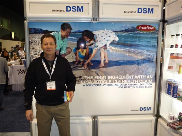 A man in a black jacket and lanyard holds two small bottles in front of a DSM display for fruitflow, showing a beach scene and text about an EFSA Article 13.5 health claim.