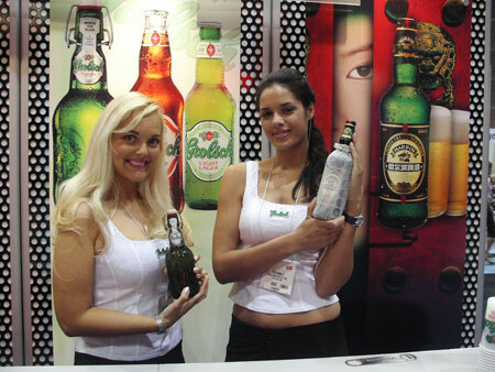 Two smiling women in white tops hold Grolsch beer bottles at a counter, with large bottle graphics of Grolsch lager behind them.