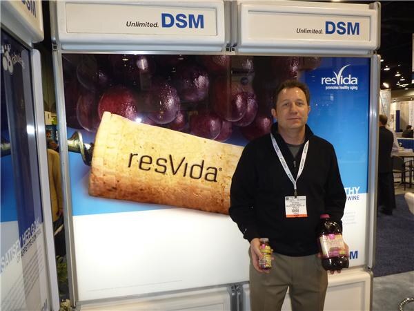 A man in a black sweater and lanyard holds two small bottles of dark purple juice in front of a DSM resVida display showing grapes and a wine cork.