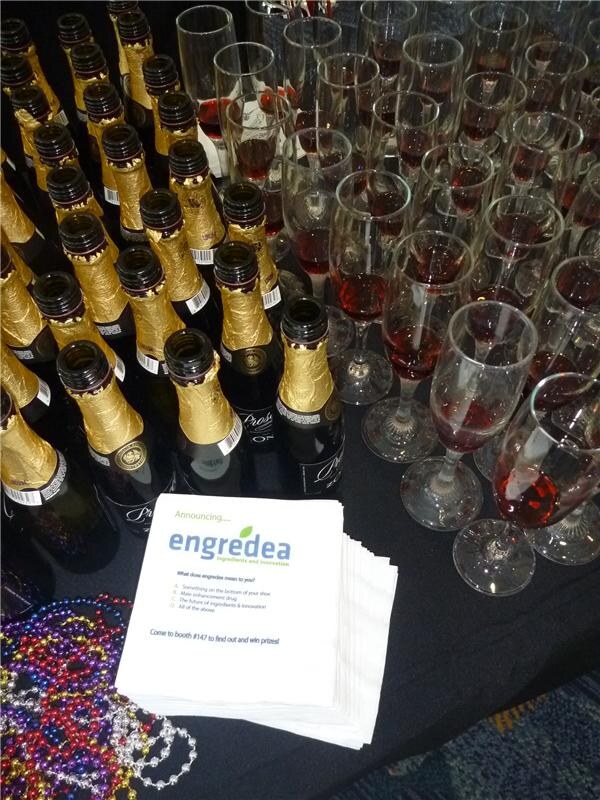 A black-draped table holds rows of gold-foil-necked bottles, glasses of red drink, colorful bead necklaces, and a stack of white napkins printed with engredea and an invitation to booth 147.