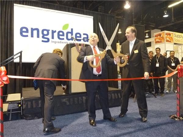 A man in a dark suit and red tie holds oversized scissors beside a red ribbon in front of an Engredea sign; a second man holds the ribbon and others stand behind.