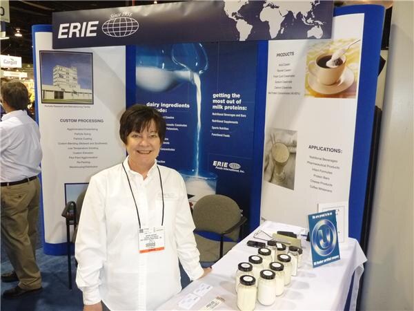 A smiling woman in a white jacket and lanyard stands at the Erie booth. Jars of white powder sit on the table, and banners describe dairy ingredients and custom processing.