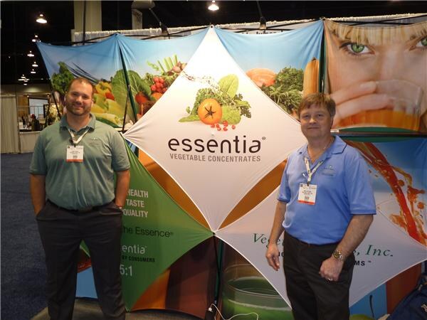 Two men stand in front of an Essentia Vegetable Concentrates display with colorful vegetable and juice graphics; one wears a gray-green polo and the other a blue polo.