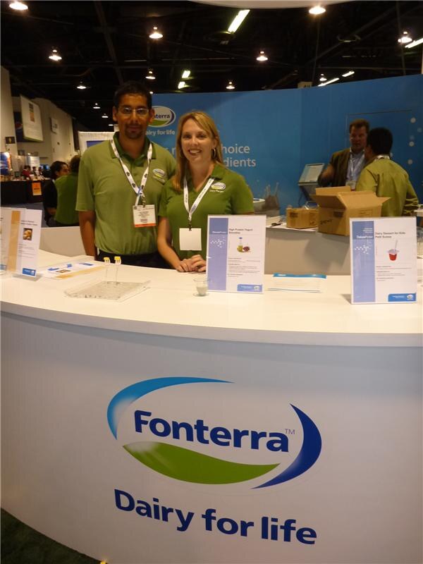 A man and a woman in green polo shirts and lanyards stand smiling behind a curved white Fonterra counter reading Dairy for life, with product information cards on top.