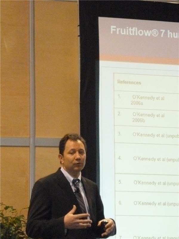 A man in a dark suit and striped tie speaks and gestures beside a projected slide titled Fruitflow with a list of references.