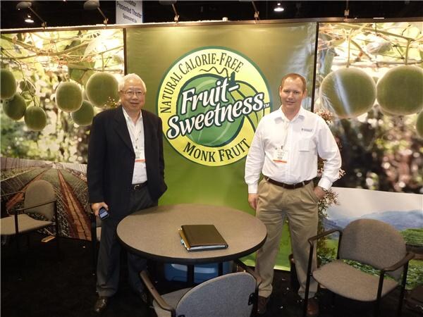 Two men stand beside a round table in front of a green Fruit Sweetness Natural Calorie-Free Monk Fruit banner. One wears a dark jacket, the other a white shirt and khaki pants.