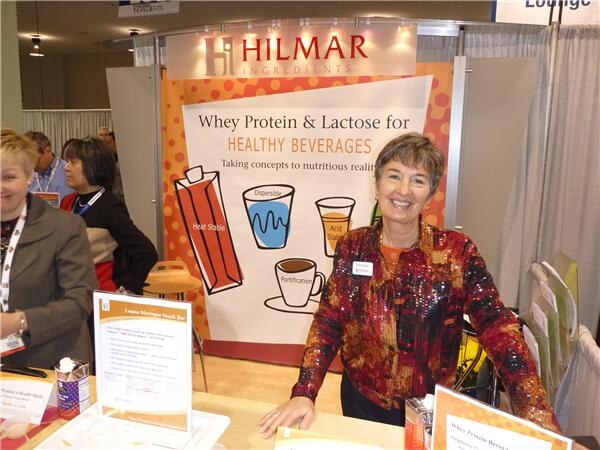 A smiling woman in a sequined jacket stands at a counter in the Hilmar Ingredients booth, in front of a sign reading Whey Protein & Lactose for Healthy Beverages. Another woman is partly visible at left.