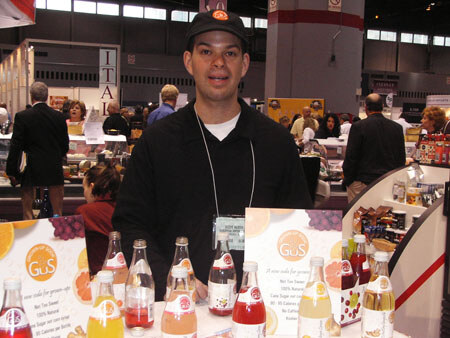 A man in a dark cap and black jacket stands behind a counter lined with GuS sparkling soda bottles, with GuS signs showing citrus and grape artwork.