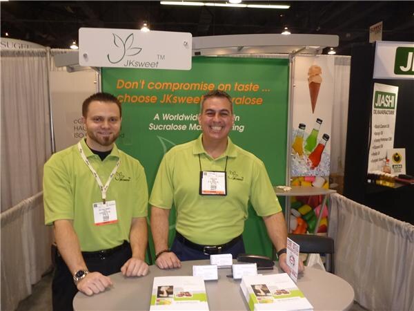 Two smiling men in light green polo shirts stand behind a table with brochures in the JKsweet booth. A green banner reads Don't compromise on taste, choose JKsweet Sucralose. A Jiashi sign is at right.