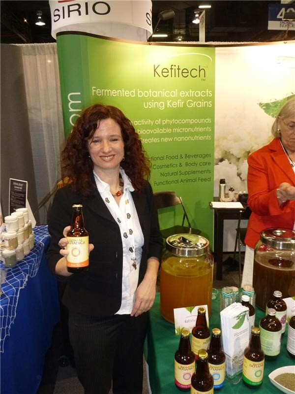 A smiling woman with long curly hair holds a Botanica bottle in front of a green Kefitech banner about fermented botanical extracts. More bottles and glass jars sit on the table, and a Sirio sign hangs above.