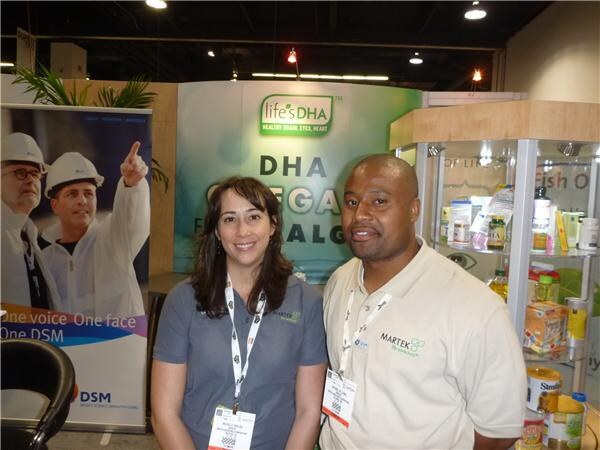 A woman and a man pose together in a booth with a life's DHA banner and a DSM banner; he wears a Martek shirt, and product bottles sit on shelves at right.